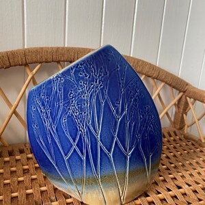 May include: A blue ceramic vase with a textured design of branches and leaves. The vase has a light brown glaze at the bottom.