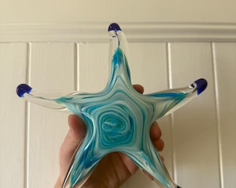 May include: A clear glass starfish with a swirling pattern of turquoise and white. The tips of the starfish's points are a deep blue. The decorative piece is held up against a white background.