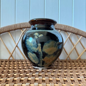 May include: A round ceramic vase with a dark brown rim and base, and a glossy black body. The vase features a unique pattern of blue and tan brushstrokes, adding an artistic touch. The vase is sitting on a woven wicker surface.
