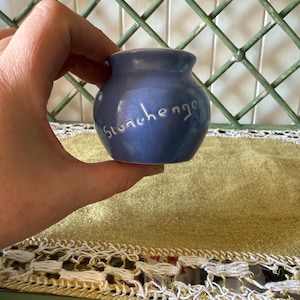 May include: A small, blue ceramic pot with the word "Stonehenge" written in cursive. The pot is held in a hand, with a green lattice background and a gold and white lace trim.