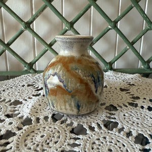 May include: A small ceramic vase with a rounded body and a narrow neck. The vase has a mix of earthy colours, including brown, beige, and blue, with a textured, glazed finish. The vase is displayed on a white, crocheted doily.