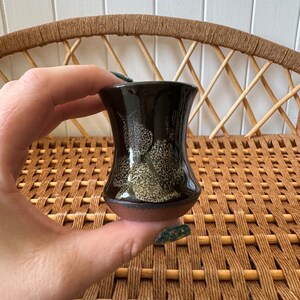 May include: A small, dark brown ceramic vase with a speckled glaze. The vase has a wide base and a narrow neck.