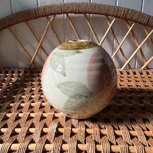 May include: A round ceramic vase with a white glaze and a pink and green floral design. The vase is sitting on a woven wicker surface.