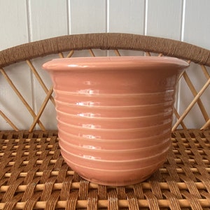 May include: A pale pink ceramic planter with a ribbed design. The planter is sitting on a woven wicker surface.