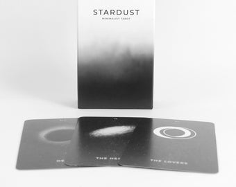 Può includere: Una scatola bianca e nera con il testo "Stardust Minimalist Tarot" sul davanti. Tre carte dei tarocchi sono disposte davanti alla scatola, mostrando le carte della Morte, dell'Eremita e degli Amanti.