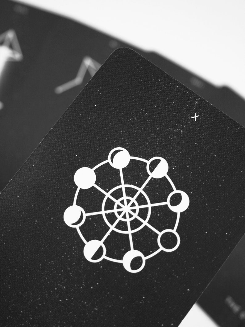 May include: Black card with a white illustration of a circle with eight moons in different phases around the edge. The card has a speckled texture.