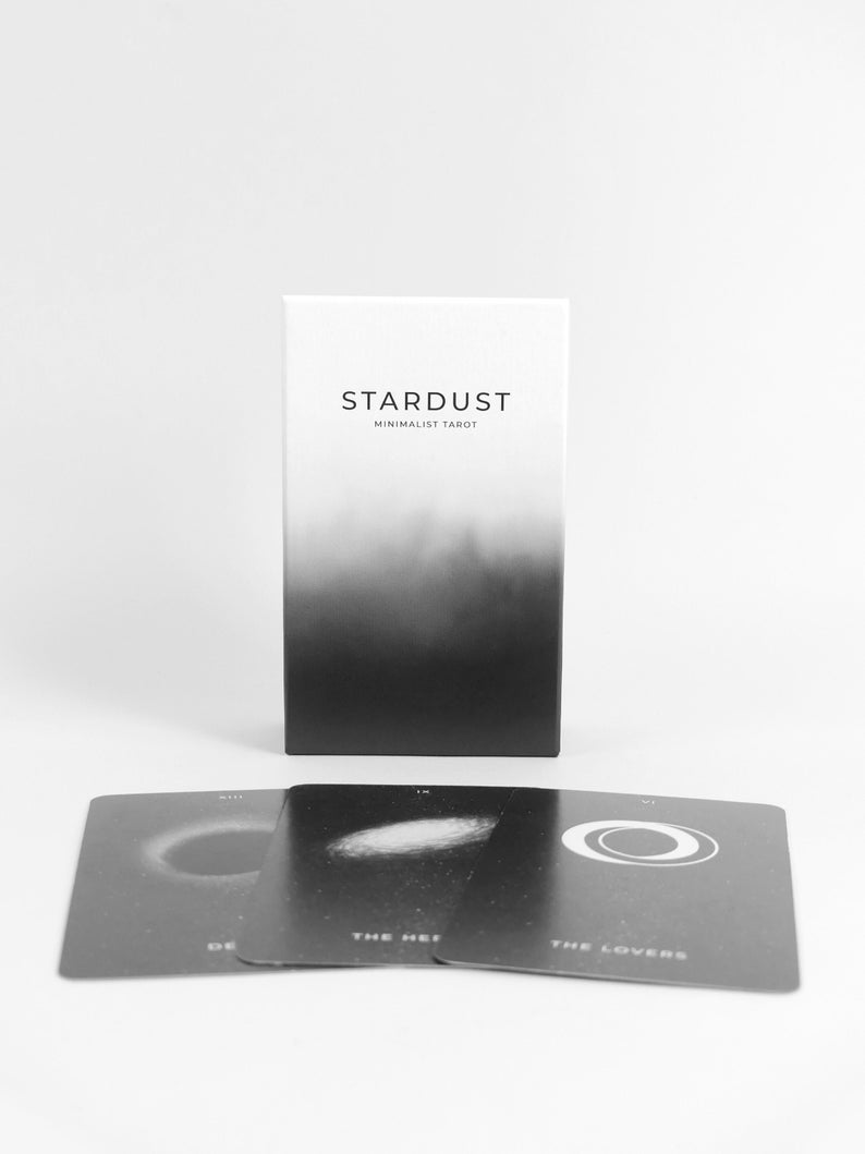 May include: A black and white box with the text "Stardust Minimalist Tarot" on the front. Three tarot cards are laid out in front of the box, showing the Death, The Hermit, and The Lovers cards.
