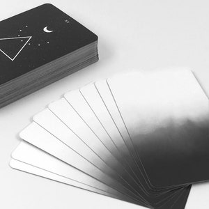May include: A deck of tarot cards with a black and white design. The cards are fanned out in a semi-circle, with the top card showing a crescent moon and a triangle with dots inside. The back of the cards are black with a white crescent moon and triangle.