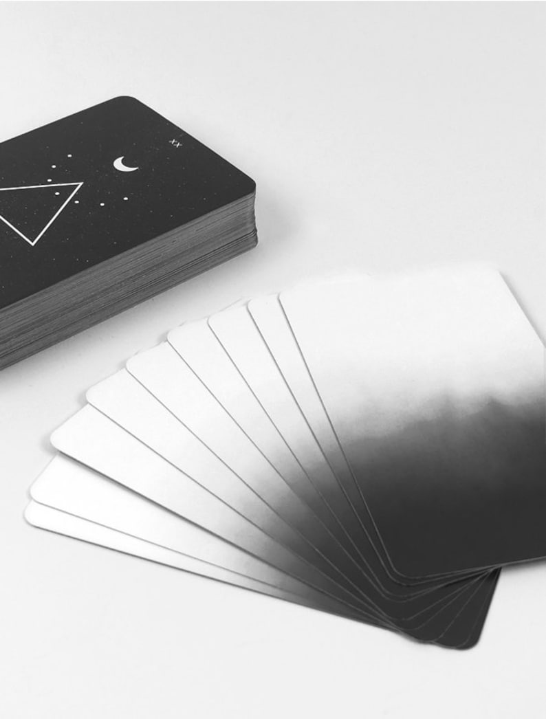 May include: A deck of tarot cards with a black and white design. The cards are fanned out in a semi-circle, with the top card showing a crescent moon and a triangle with dots inside. The back of the cards are black with a white crescent moon and triangle.