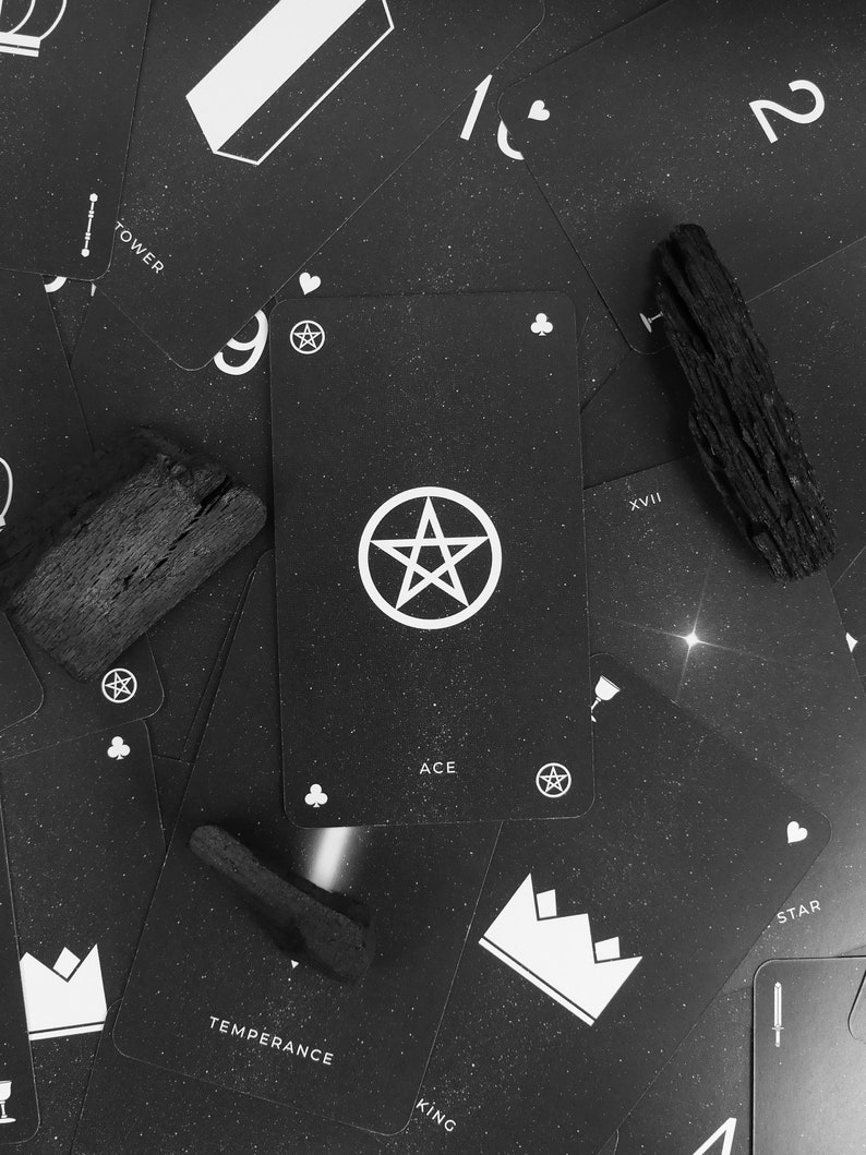 May include: Black and white tarot cards with a pentagram design on the Ace of Clubs card. The cards are scattered on a black surface with wooden sticks.