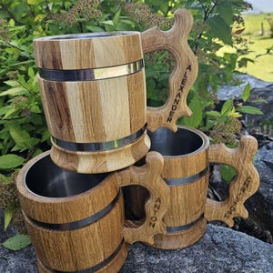 Könnte beinhalten: Drei Bierkrüge aus Holz mit Edelstahlbändern und personalisierten Namen. Die Krüge sind übereinander gestapelt. Die Namen auf den Krügen sind Alexander, Anton und True North.