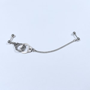 May include: A silver chain connects two silver barbell earrings with ball ends. One earring features a pair of silver handcuffs.