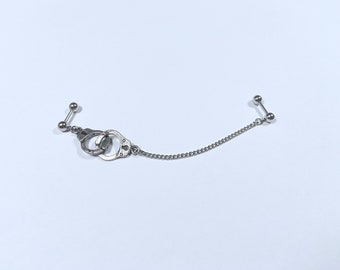 May include: A silver chain connects two silver barbell earrings with ball ends. One earring features a pair of silver handcuffs.