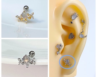 May include: A close-up of a silver and gold cartilage earring with a clear stone and a star design. The earring is shown on a white background and on a model's ear.