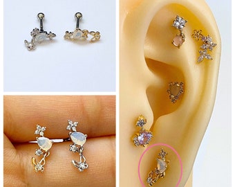 May include: A pair of silver and gold cartilage earrings with a floral design and a small opal stone. The earrings are shown on a model's ear.