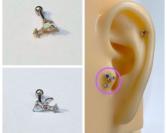 May include: A silver barbell with a clear iridescent butterfly charm and clear crystal accents. The barbell is shown in a close-up image and also worn in a person's ear.
