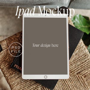 Peut inclure: Une maquette d'iPad blanche avec un écran gris affichant le texte "Your design here". L'iPad est posé sur un carnet noir et une surface en osier brun.