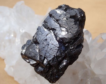 May include: A dark gray, metallic, rough-textured mineral specimen. The mineral is sitting on a bed of white quartz crystals.