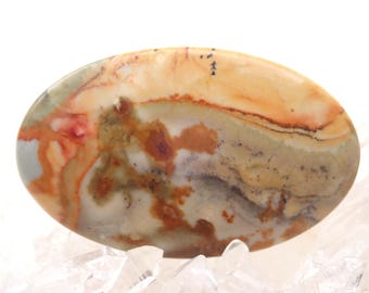 Pode incluir: Uma pedra preciosa polida de formato oval com uma superfície lisa e padrões em espiral nas cores creme, castanho, laranja queimado e verde sálvia suave. A pedra apresenta inclusões minerais naturais e gradientes de cor orgânicos.