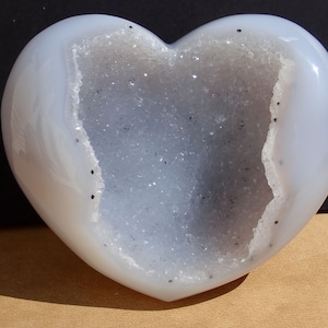 May include: A heart-shaped geode, featuring a pale blue, polished exterior and a crystalline interior. The cavity is filled with sparkling crystals, contrasting with the smooth outer surface. Small black specks are scattered across the geode's surface.