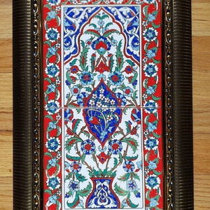 May include: A framed tile with a traditional Turkish design. The tile features a floral pattern in shades of blue, red, and green on a white background. The frame is gold with a decorative border.