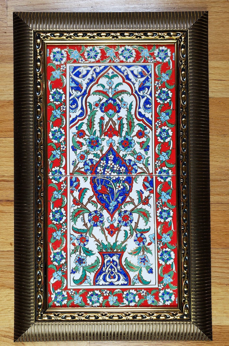 May include: A framed tile with a traditional Turkish design. The tile features a floral pattern in shades of blue, red, and green on a white background. The frame is gold with a decorative border.