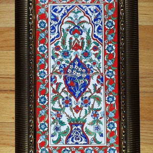 May include: A framed ceramic tile with a traditional Turkish design. The tile features a floral pattern in shades of blue, red, and green, with a white background. The tile is framed in a gold-colored frame.