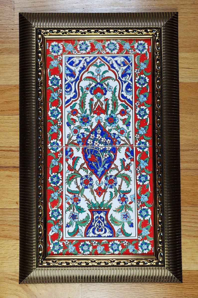 May include: A framed ceramic tile with a traditional Turkish design. The tile features a floral pattern in shades of blue, red, and green, with a white background. The tile is framed in a gold-colored frame.