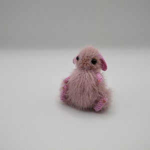 May include: A small, pink crocheted bunny with black eyes and a fluffy texture. The bunny is sitting on a white surface.
