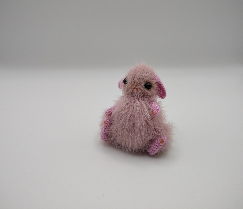 May include: A small, pink crocheted bunny with black eyes and a fluffy texture. The bunny is sitting on a white surface.