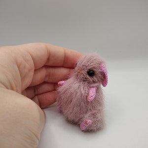 May include: A small, pink crocheted bunny with black eyes. The bunny is sitting on a white surface.