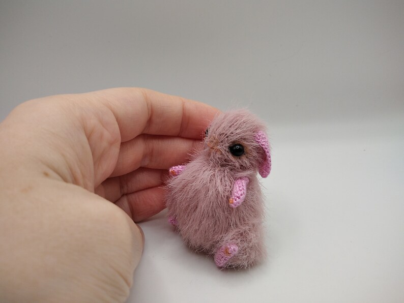 May include: A small, pink crocheted bunny with black eyes. The bunny is sitting on a white surface.