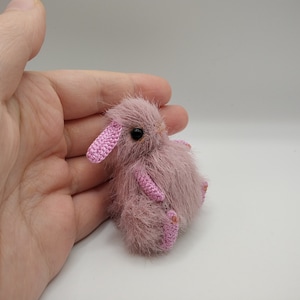 May include: A small, pink crocheted bunny with black eyes. The bunny is sitting on a hand.