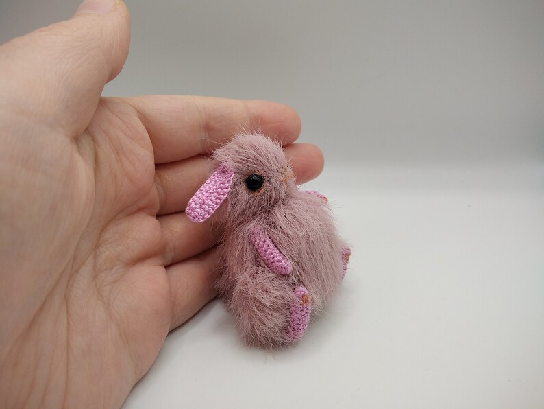May include: A small, pink crocheted bunny with black eyes. The bunny is sitting on a hand.