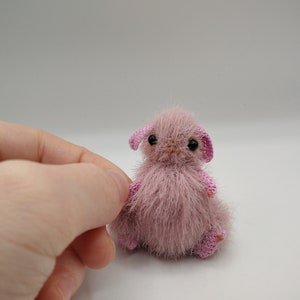 May include: A small, pink, crocheted animal with black eyes and pink ears. The animal is sitting on a white surface.