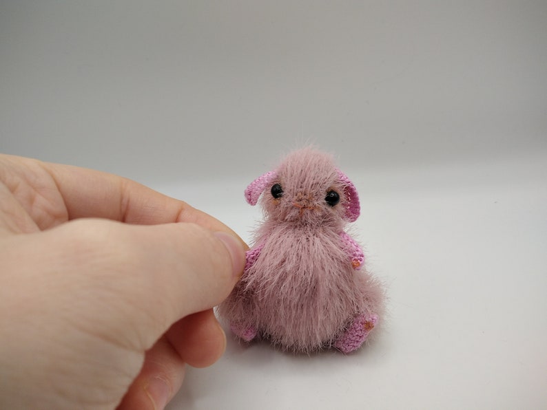 May include: A small, pink, crocheted animal with black eyes and pink ears. The animal is sitting on a white surface.