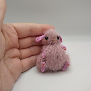 May include: A small, pink, crocheted bunny with black eyes and pink ears. The bunny is sitting on a white surface.
