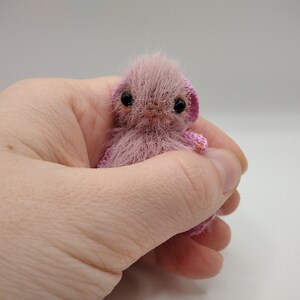 May include: A small, pink crocheted stuffed animal with black eyes and a pink nose. The animal is sitting in a hand.