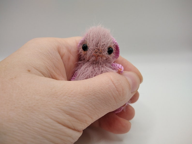 May include: A small, pink crocheted stuffed animal with black eyes and a pink nose. The animal is sitting in a hand.