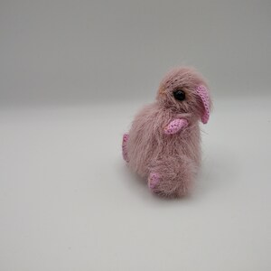 May include: A small, pink crocheted bunny with pink ears and black eyes. The bunny is sitting on a white surface.