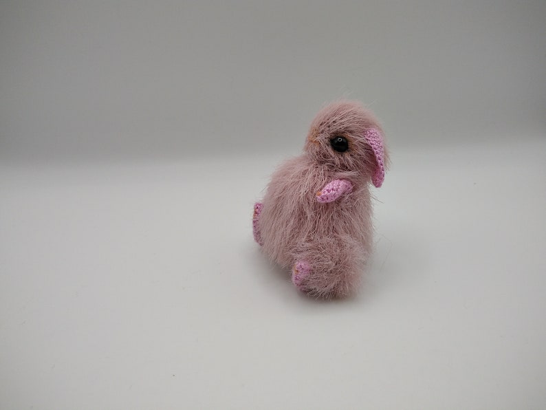 May include: A small, pink crocheted bunny with pink ears and black eyes. The bunny is sitting on a white surface.