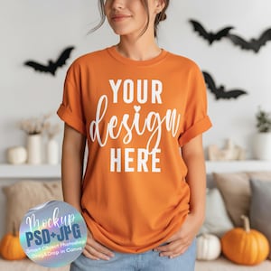 Puede incluir: Camiseta naranja con mangas enrolladas que muestra el texto blanco "YOUR design HERE". La imagen es una maqueta, con decoraciones de Halloween en el fondo, incluyendo murciélagos y calabazas.