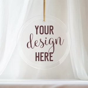 Può includere: Un ornamento rotondo in vetro trasparente con un filo dorato appeso in cima. L'ornamento ha il testo "YOUR design HERE" stampato in marrone scuro.