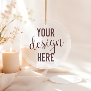 Puede incluir: Un adorno redondo de acrílico transparente con el texto "YOUR design HERE" colgado de una cuerda dorada. El adorno está delante de un fondo blanco con una vela y flores blancas.