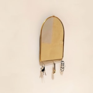 Könnte beinhalten: Ein goldener Bogenspiegel mit einem Schlüsselhalter, der an der Unterseite befestigt ist. Der Schlüsselhalter hat drei Haken und eine Kette, die daran hängt.