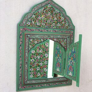 May include: A green wooden mirror with intricate floral designs painted on the frame. The mirror has two doors that open to reveal a smaller mirror inside.