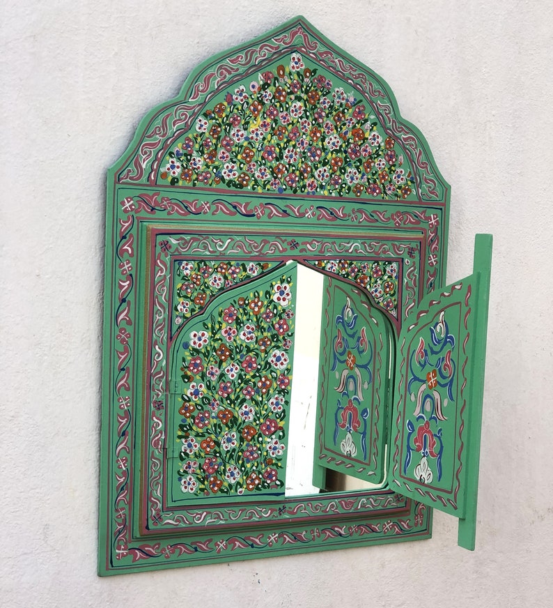 May include: A green wooden mirror with intricate floral designs painted on the frame. The mirror has two doors that open to reveal a smaller mirror inside.