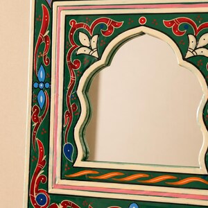 Pu&ograve; includere: Specchio decorativo con cornice dipinta di verde, rosso e bianco. La cornice presenta un'apertura ad arco e intricati disegni floreali e geometrici. Il bordo dello specchio include accenti rosa e blu.