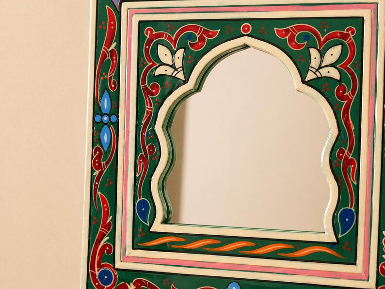 Pu&ograve; includere: Specchio decorativo con cornice dipinta di verde, rosso e bianco. La cornice presenta un'apertura ad arco e intricati disegni floreali e geometrici. Il bordo dello specchio include accenti rosa e blu.