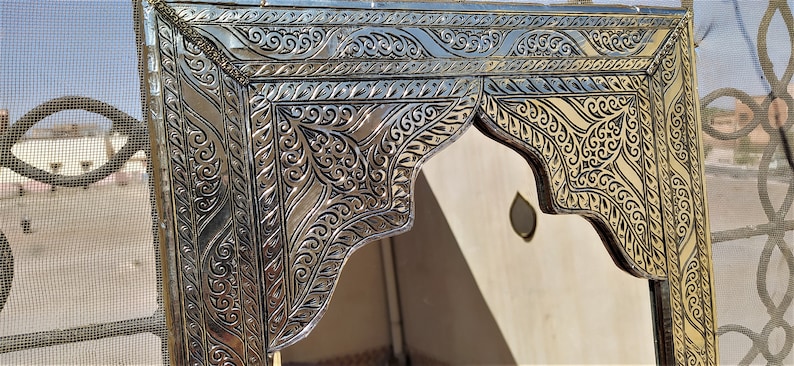 May include: A silver and gold ornate mirror with intricate floral designs. The mirror has a rectangular frame with a decorative arch at the top.